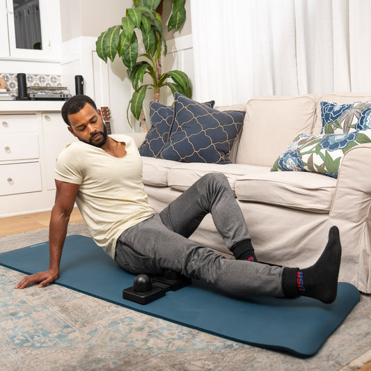 man in his home using Njoie's MFLEX myofascial release deep tissue massage tool on his hamstrings, upper legs, back of legs, glutes
